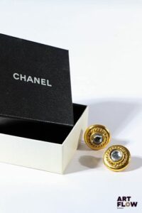CHANEL Paire de clips d'oreilles en métal doré de forme ronde centrée chacune d'un strass. Diamètre : 2,5 cm environ Dans leur boîte d'origine.