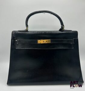 HERMÈS Paris Circa 1950 Sac à main Kelly en cuir box noir, garnitures en métal doré, avec cadenas et clochette avec clés. Intérieur à une poche plaquée double à l'avant et une poche plaquée simple à l'arrière du sac. H : 25 x L : 32 x P : 13 cm Usures, défauts d'usage, craquelures, griffures et frottements Dustbag