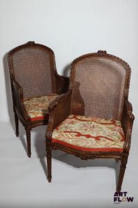 Paire de larges bergères cannées en bois mouluré et sculpté, accotoirs à manchette. Garnies d'un carreau amovible de tapisserie au petit point. De style Transition. 99 x 64 x 45 cm. Accidents et restaurations.
