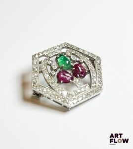 Broche en platine et épingle en or blanc 750‰ (poinçons tête d’aigle et tête de chien), de forme hexagonale, en forme de bouquet de fleurs dans un cadre géométrique, délicatement ajouré, entièrement pavé de diamants taille ancienne ; le bouquet représenté par une émeraude poire cabochon et deux rubis sculptés dont un percé. Bouquet dessoudé sur deux points à l’arrière. Poids : 7.16 g brut Dimensions : 2.4 x 2.9 cm Expertise : Philippine Vanheems
