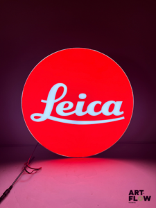 Enseigne lumineuse publicitaire sur plexiglas Leica. Dimensions : 53 x 35 cm Fonctionnelle - État général excellent. Fourni avec son alimentation murale.
