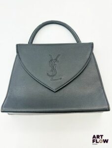YVES SAINT LAURENT Sac à main en cuir grainé noir monogrammé "YSL" en point de couture, fermeture bouton-pression, une poche extérieure, intérieur avec deux poches dont l'une se ferme à l'aide d'un zip. Dimensions : 28 x 21 x 7 cm Quelques marques d'usure sur le cuir, anse décollée au niveau de la jonction avec le sac et marques d'usures, intérieur en bon état.